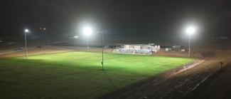Photo of large grass rectangular field with flood lights in each corner and a building on the edge with tiered seating in front of it.