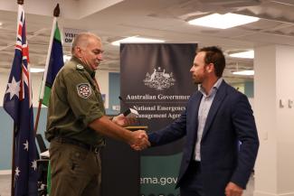 Tony Fuller in a khaki-coloured uniform with an insignia patch on his sleeve shaking hands with Dave Long. They are standing in front of a banner with the Australian Government crest that says National Emergency Management Agency.