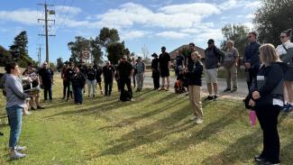 Photo of a group of people standing around on a grassy area beside a road, listening to someone speak.