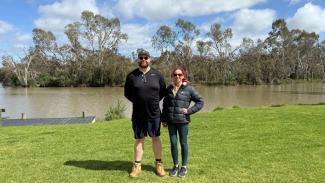 Photo of two people standing next to each other looking at the camera, on a grassy area with a river in the background and gumtrees on the far shore.