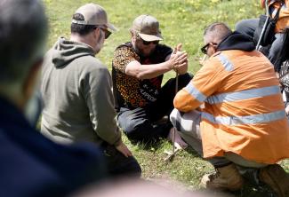 Photo of a group of people crouching and sitting around a man starting a fire with sticks.