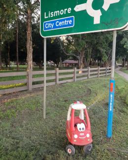 Photo of a toddler toy ride-in firetruck with big eyes and a smile, sitting under a road sign to Lismore.