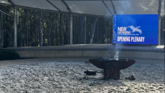 Photo of a sandy area with a bench seat around the edge, a smouldering fire pit in the centre, and a screen behind that says NIDR Gathering Opening Ceremony.