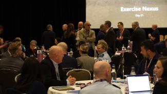 Photo of the conference with groups of people sitting at round tables, some with laptops in front of them, and some people standing up and mingling in the background. A large screen at the back has Exercise Nexus Day 1 on it.