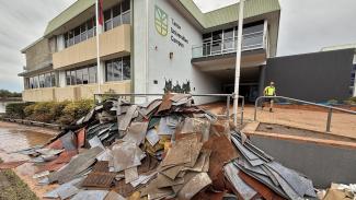Photo looking up at a building with Taree Universities Campus sign and logo on the side, with a very large pile of rubble stacked up in front, mostly containing sheets of carpet and metal.