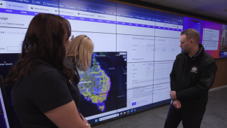 Photo of ICG Pat Hetherington in front of floor to ceiling screens displaying a map of Australia and text fields. He is talking to 2 other people, looking at the screens.