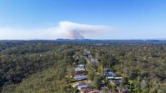 Aerial photo of dense bush with some houses in the middle and in the far distance on the horizon, bushfire smoke is rising into a blue sky.