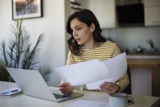 Woman sitting at a table looking slightly concerned at a laptop with papers in hand.