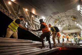 Group of people wearing high vis vests inside the hull of an aeroplane, unloading long pieces of metal and other cargo.