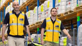 Two men carrying a generator between them, wearing high vis yellow vests, walking in a warehouse with tall stacked shelves behind them of packages labelled 'Australian Aid'.