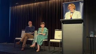 Photo of a woman speaking at a lectern and a man and a woman sitting on the stage next to her watching her present.
