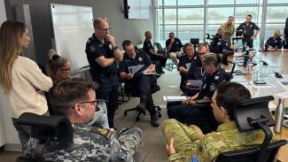 Photo of a group of people in chairs and standing, not around a table, whiteboard in background, some people are wearing Defence uniforms.