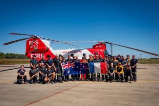 Large group of people posing in front of a large red and white RFS helicopter, holding large Australian and French flags, on a concrete runway area with blue sky.
