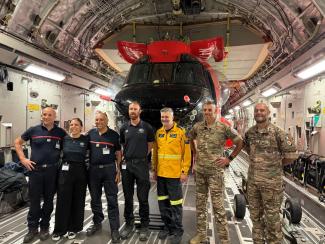 Seven people standing in a row, some in NEMA uniform, some in French Army uniform and 1 in yellow firefighter uniform. They are standing in front of a red helicopter, all inside a very large aircraft with cables and pipes visible across the curved ceiling.